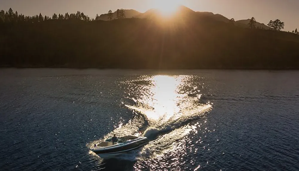 A boat speeds across a calm lake with the sun setting behind a mountainous backdrop