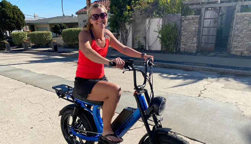 A person is smiling while sitting on a blue electric bicycle on a sunny day in a residential area