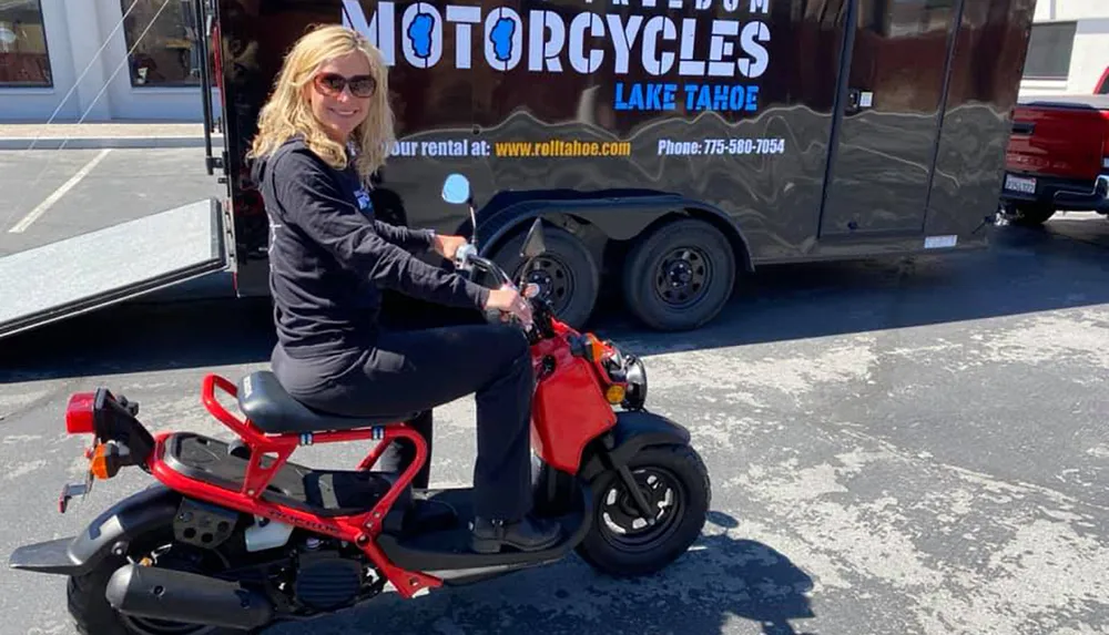 A smiling person is seated on a red and black mini motorcycle in a parking lot with a vehicle sporting a Motorcycles Lake Tahoe advertisement in the background