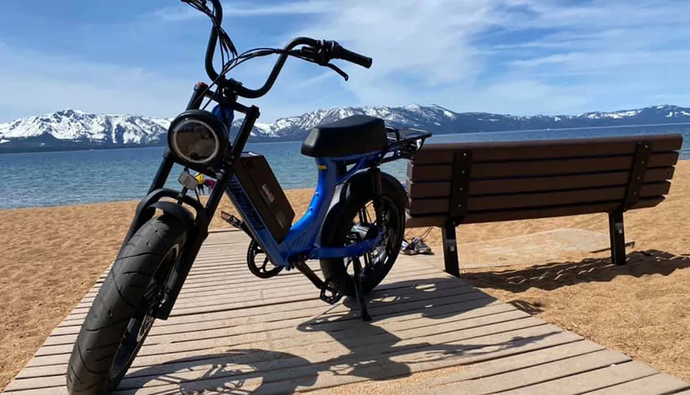 A blue electric bicycle is parked on a wooden boardwalk by a sandy beach with a backdrop of snowy mountains and a clear blue sky