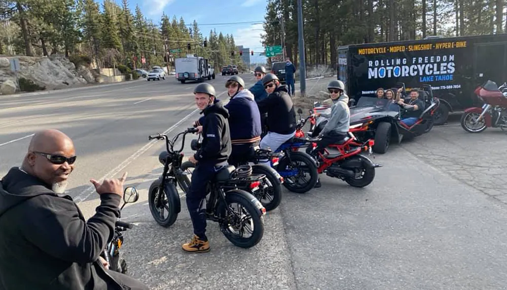 A group of people with a mix of motorcycles and an open-cockpit vehicle are lined up on a roadside smiling and posing for the photo