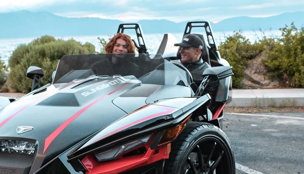 Two individuals are enjoying a ride in a Polaris Slingshot against a backdrop of nature