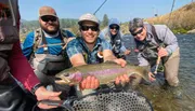 A group of smiling people are standing in a river with fishing gear, as one of them holds a large trout for the camera.