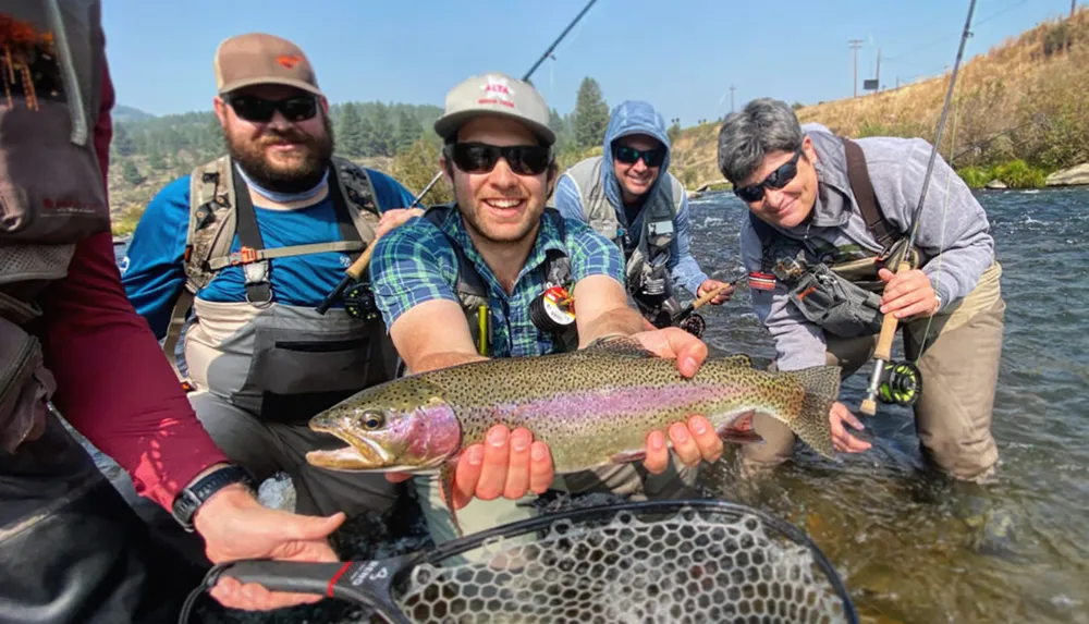 A group of smiling people are standing in a river with fishing gear as one of them holds a large trout for the camera