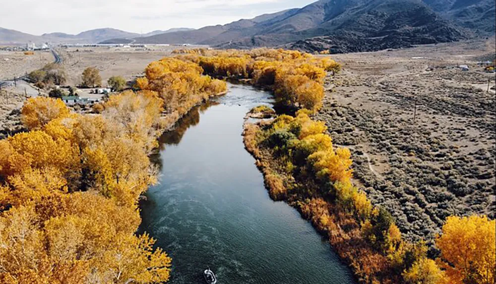 A winding river bordered by trees with autumn foliage meanders through a sparse landscape with a small boat visible on the water