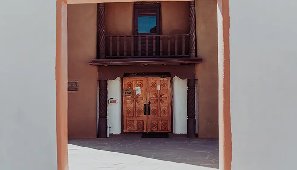 The image shows a traditional Pueblo-style entrance with a carved wooden door and a small upper balcony framed by a terra cotta-colored archway