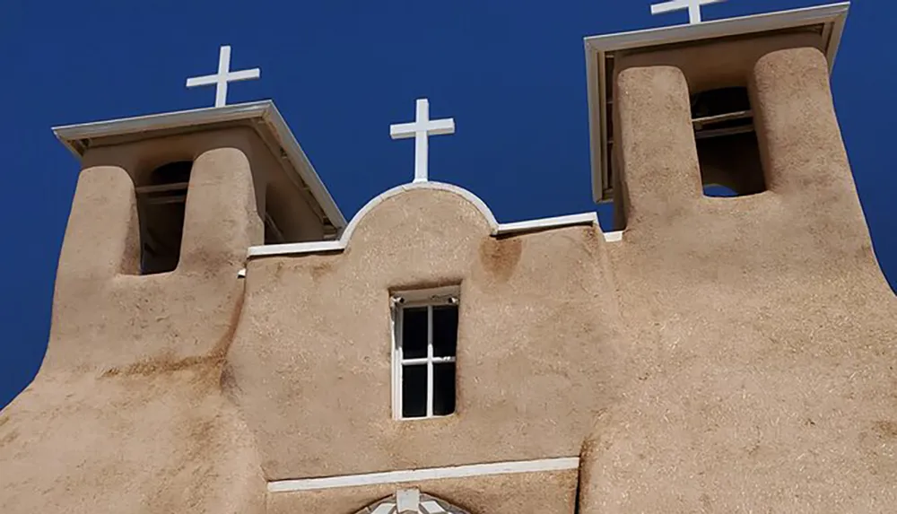 The image shows the upper part of an adobe church with twin bell towers topped with crosses against a blue sky