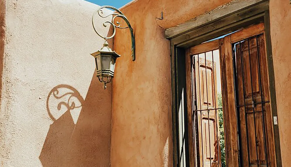 An ornate street lamp casts a decorative shadow on a sunlit warm-colored adobe wall next to a wooden door