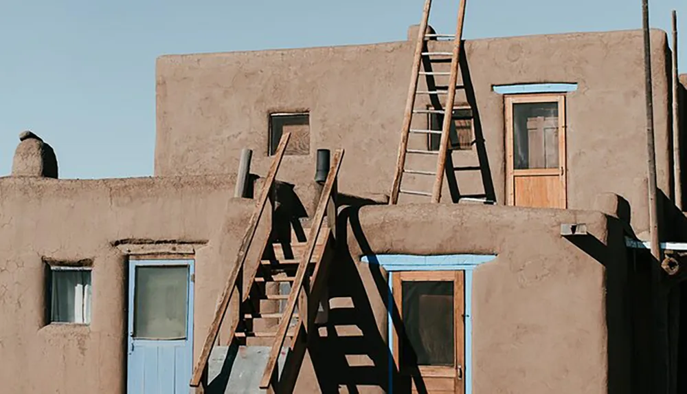 The image shows a section of a traditional adobe building with wooden ladders extending to its flat roof against a clear blue sky