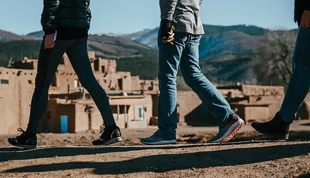 Two people are walking past each other on a dirt path with traditional adobe buildings and a hilly landscape in the background