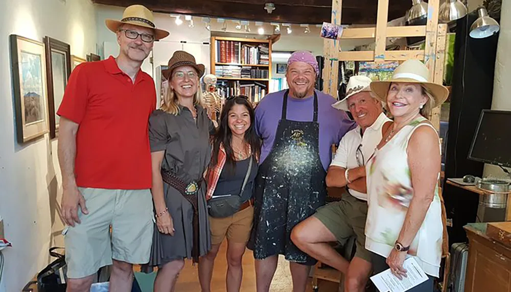 A group of six smiling people some wearing hats pose together inside a room with artwork and books exhibiting a casual and joyful atmosphere