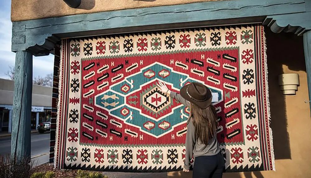 A person wearing a hat is admiring a large colorful rug with intricate patterns hung on the exterior wall of a building