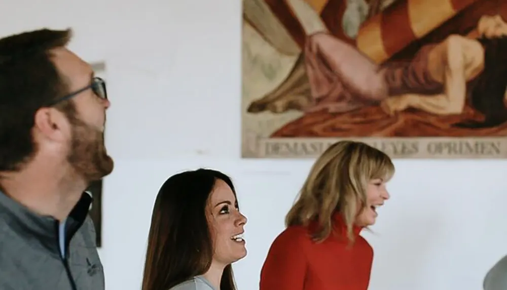 Three people appear to be engaged in conversation or looking at something out of frame with a piece of art displaying text that suggests a theme of storytelling or history on the wall behind them