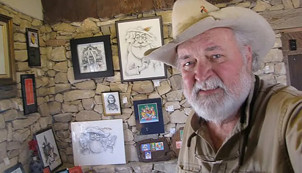 A man in a cowboy hat smiles for a selfie in a room adorned with various framed artworks on a stone wall