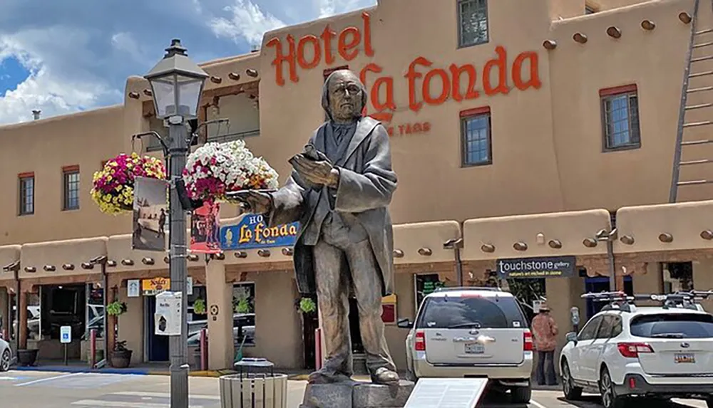 A statue of a seated figure holding a bird stands in front of the Hotel La Fonda with parked cars and a flower-adorned lamp post under a blue sky with fluffy clouds