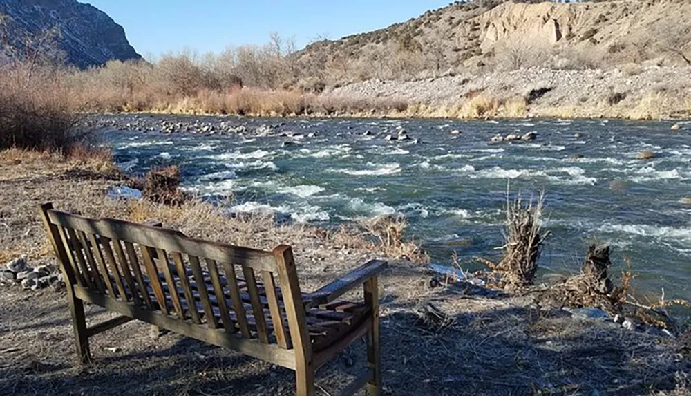 A solitary wooden bench sits facing a rugged rippling river flanked by dry vegetation and a rocky landscape under a clear sky
