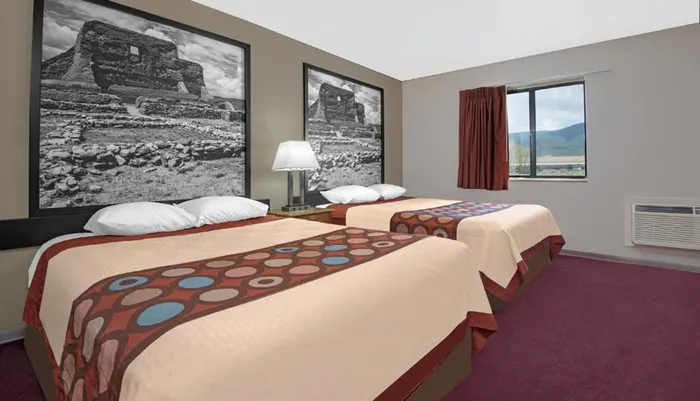 The image shows a hotel room with two beds featuring patterned bedspreads a large black-and-white landscape photograph on the wall and a window with a view of the mountains