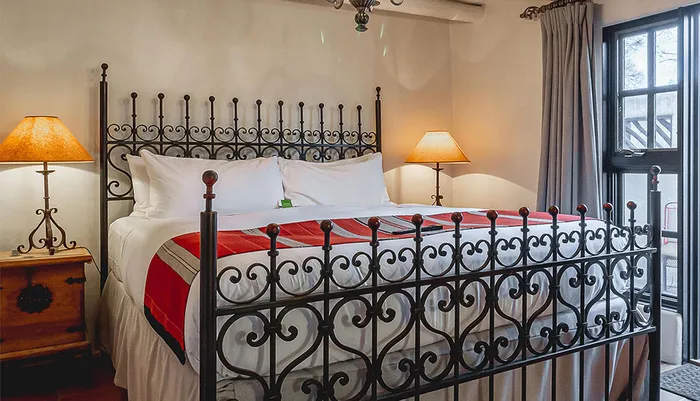 The image shows a neatly-made bed with a wrought iron headboard flanked by two bedside tables with lamps in a cozy room with a window