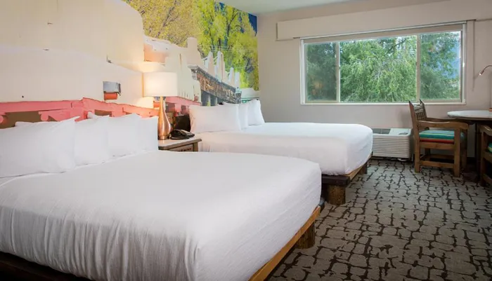 The image shows a bright hotel room with two queen-sized beds a nature-themed mural behind the headboard a window with a tree view and a desk with a chair