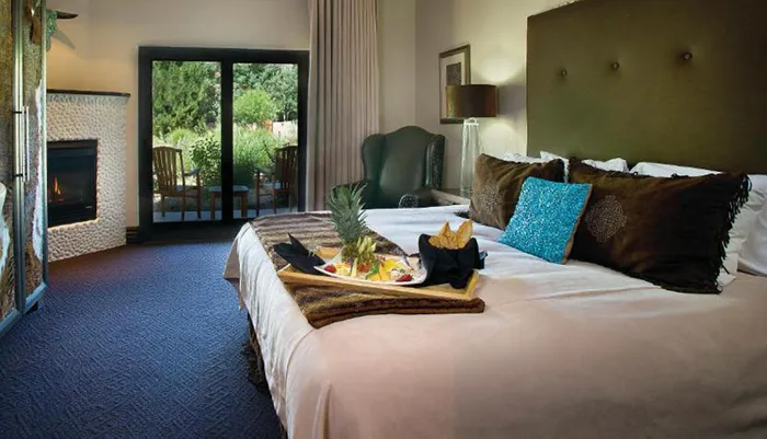 The image shows a well-appointed hotel room with a tray of food on the bed a fireplace to the left and an open glass door leading to an outdoor seating area