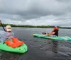 Two people are kayaking on calm waters with overcast skies one in a green kayak and another in a yellow and blue one both wearing life vests