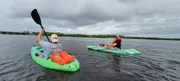 Two people are kayaking on calm waters with overcast skies, one in a green kayak and another in a yellow and blue one, both wearing life vests.
