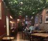 The image shows a cozy restaurant interior with people dining under a canopy of greenery and string lights with brick walls adorned with framed pictures