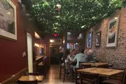 The image shows a cozy restaurant interior with people dining under a canopy of greenery and string lights, with brick walls adorned with framed pictures.