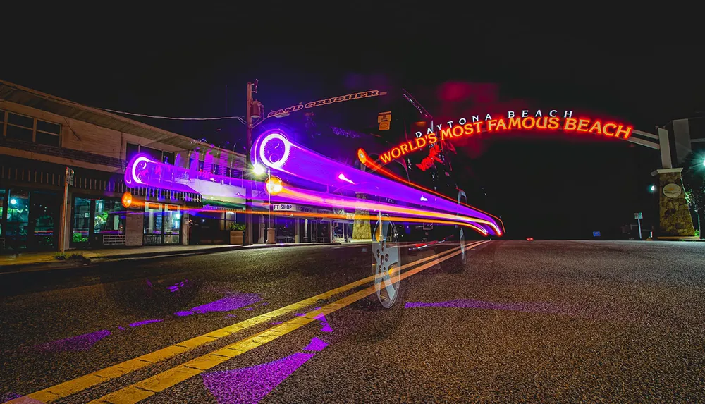A long exposure photograph captures the light trails of a passing vehicle beneath the illuminated archway sign that reads Daytona BeachWorlds Most Famous Beach