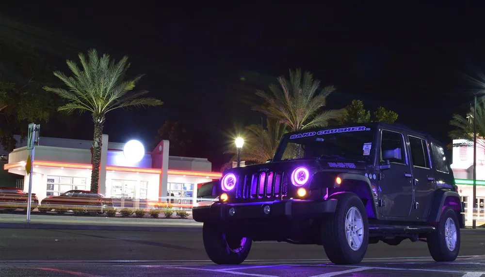 A black Jeep with purple headlights is parked at night with palm trees and a brightly lit building in the background