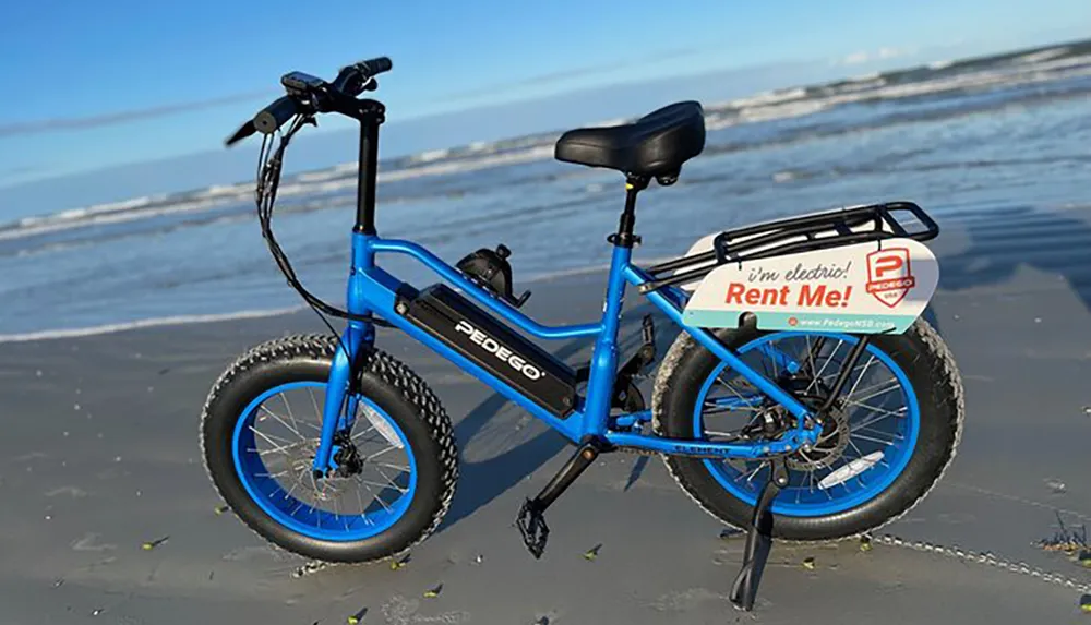 A blue electric fat-tire bike with a Rent Me sign is parked on a sandy beach with the ocean in the background