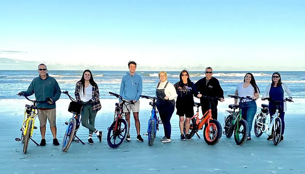 A group of seven people is standing with bicycles on a beach smiling for the camera with the ocean and a clear sky in the background