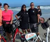 Four people are smiling for a photo on a sandy beach with bicycles two of which have signs saying Rent Me indicating they may be rental bikes