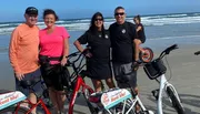 Four people are smiling for a photo on a sandy beach with bicycles, two of which have signs saying 