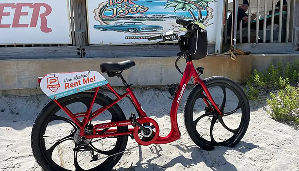 A red electric rental bicycle is parked on a sandy area in front of a sign advertising beer and the presence of a casual eatery called Sloppy Joes with a person sitting in the background