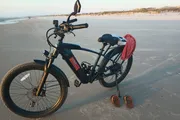 An electric bicycle is parked on a sandy beach with a pair of flip-flops placed beside it as the sun sets, suggesting a leisurely pause in someone's ride.