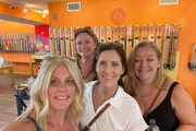 Four women are posing for a selfie with smiles, in a store with colorful wall-mounted candy dispensers in the background.