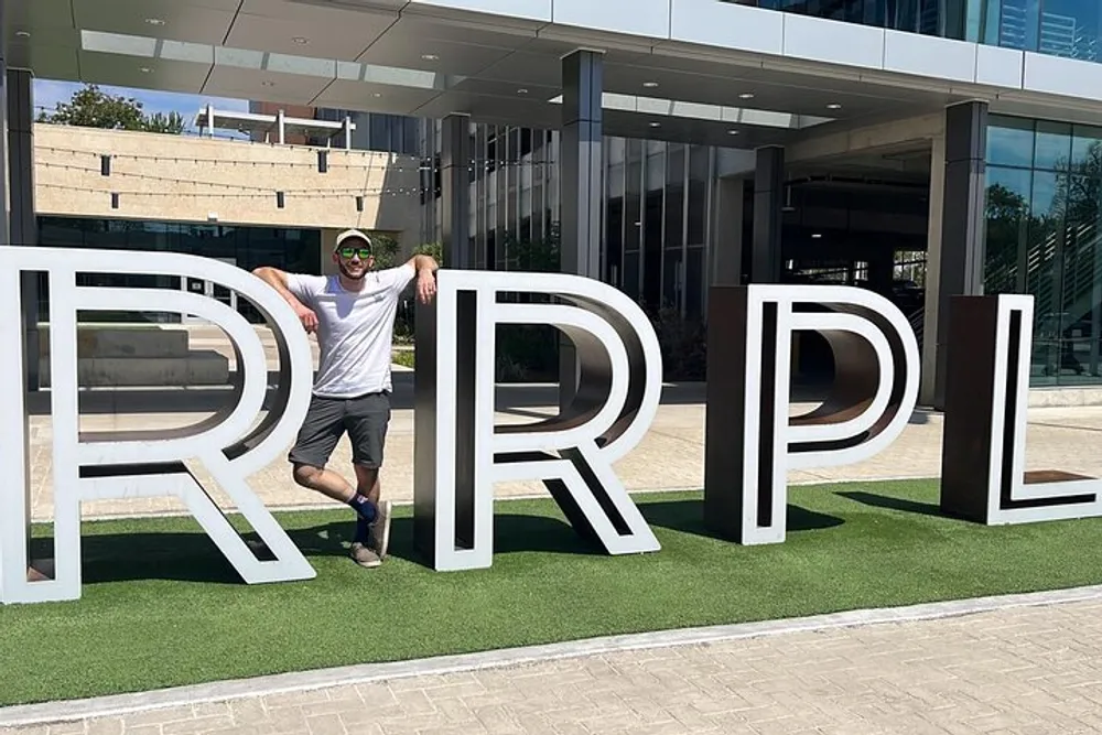 A man in casual attire is posing next to oversized letters RRPL installed on a grassy area in front of a modern building
