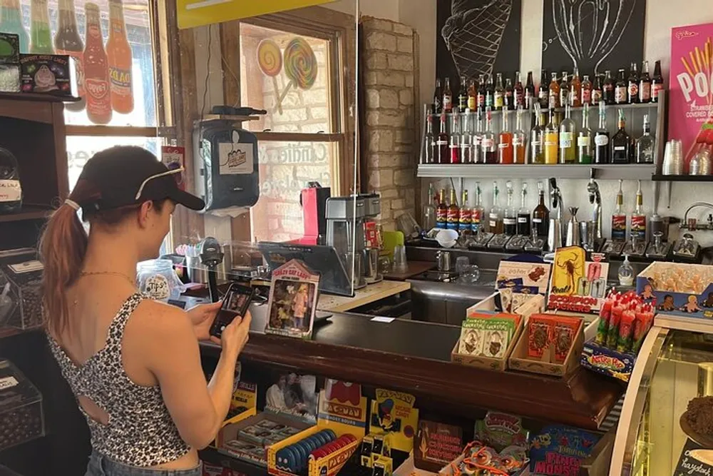 A person is using a mobile device behind the counter of a colorful and eclectic shop filled with various beverages and snacks