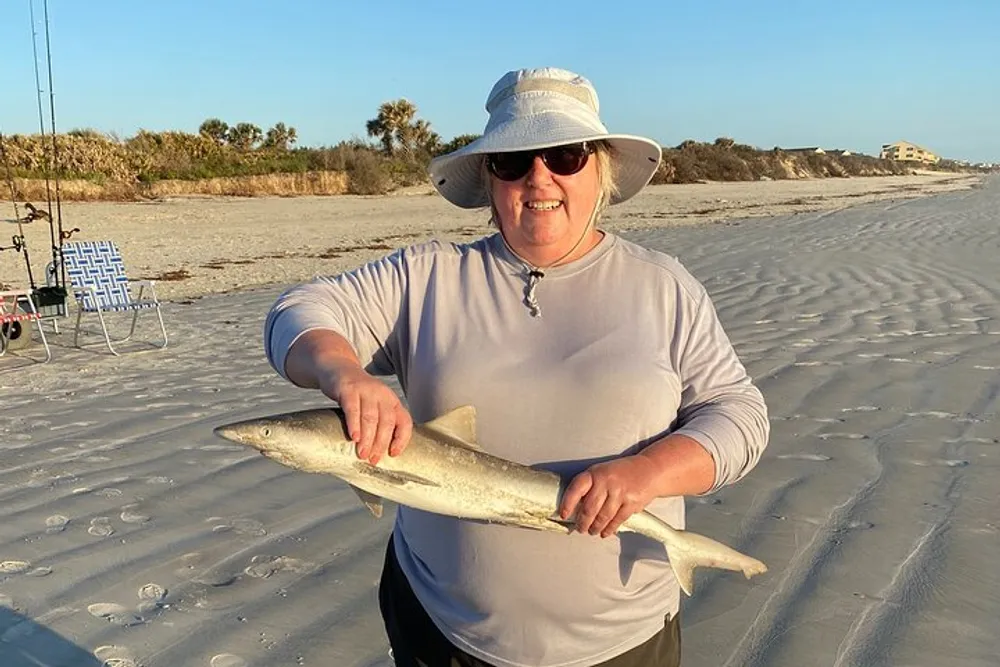 A person is smiling for the camera while holding a small shark on a sandy beach with fishing equipment in the background during what appears to be late afternoon