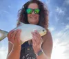 A smiling woman wearing sunglasses holds up a fish to the camera against a bright sky backdrop