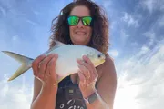 A smiling woman wearing sunglasses holds up a fish to the camera against a bright sky backdrop.