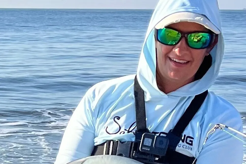 A person wearing sunglasses and protective outdoor clothing is smiling on a boat with the ocean in the background