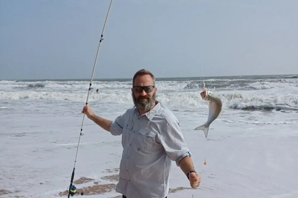 A man is holding a fishing rod and showing off a fish he caught on a sandy beach with waves in the background