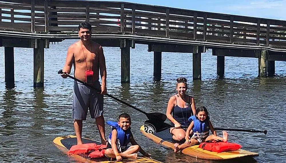 A family of four is enjoying time together paddle boarding near a wooden pier on a sunny day
