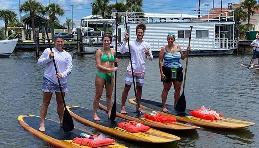 Four individuals are smiling and posing on stand-up paddleboards on a sunny day with houseboats and docks in the background