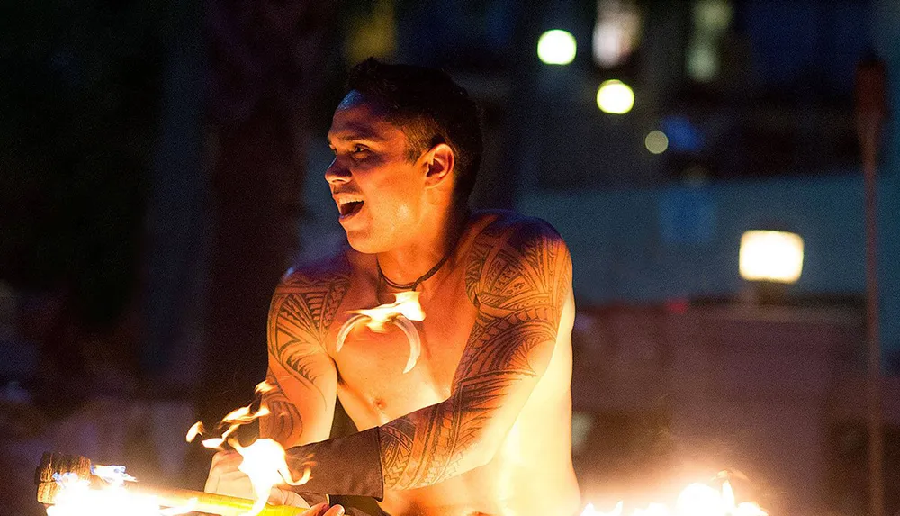 A shirtless performer with traditional tattoos is handling fire in a night-time setting exuding confidence and focus