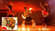 Performers are engaged in a dynamic fire dance, possibly part of a cultural performance, with a man in the center twirling fire sticks against a dramatic, fiery backdrop.
