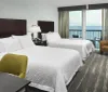 The image shows a neat hotel room with two beds and a balcony overlooking the ocean