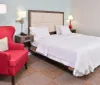The image shows a neatly made king-sized bed with white bedding in a tidy hotel room accompanied by a red accent chair and contrasting modern lamps